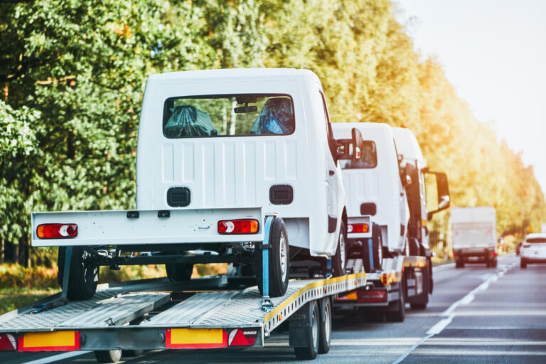 Ciężarówka z płaską platformą przewozi małe białe samochody dostawcze po autostradzie, otoczonej drzewami i innymi pojazdami pod czystym niebem.
