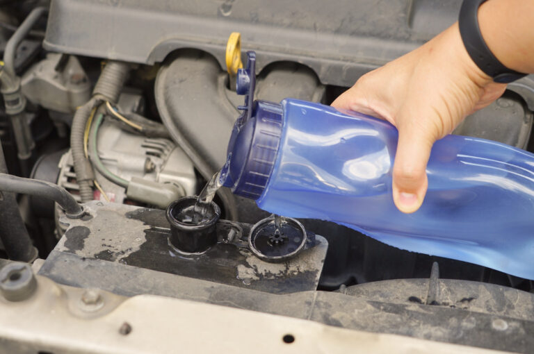 Ręka wlewa wodę z niebieskiej butelki do zbiornika chłodnicy samochodu pod maską, przy czym woda rozlewa się wokół otworu.