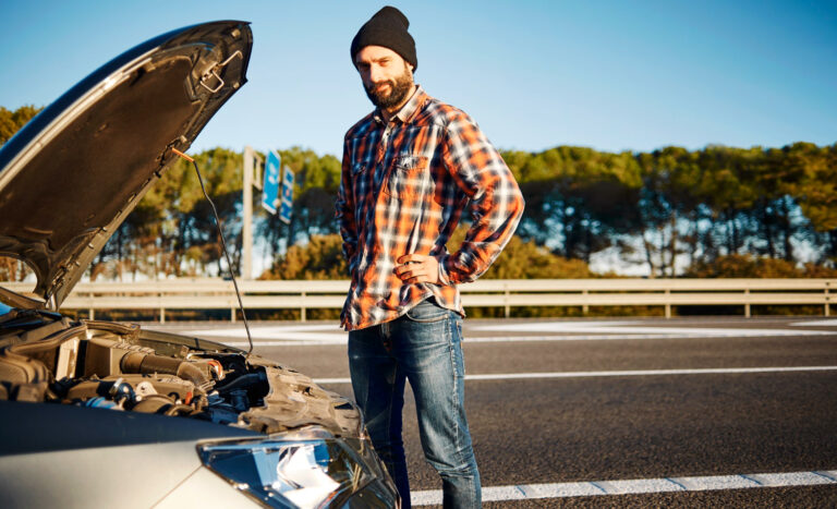 Mężczyzna w kraciastej koszuli i czapce stoi obok samochodu z otwartą maską na poboczu drogi, wyglądając na zaniepokojonego. Drzewa i barierka na autostradzie są widoczne w tle pod czystym niebem.