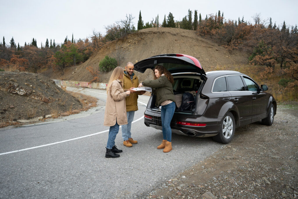Trzy osoby stoją przy otwartym bagażniku SUV-a na poboczu drogi, dzieląc się pizzą na świeżym powietrzu. Dwie kobiety i jeden mężczyzna są uśmiechnięci i ciepło ubrani; w tle widać wzgórza i drzewa.