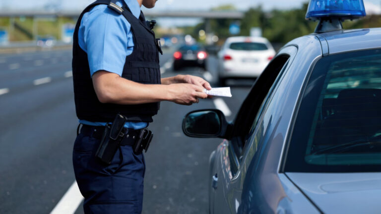 Policjant w mundurze stoi obok samochodu na autostradzie, trzymając kartkę papieru i rozmawiając z kierowcą przez okno. W tle widać kilka samochodów.