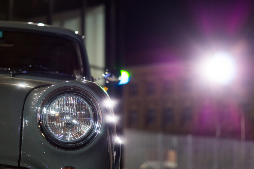 Zbliżenie przedniego lewego reflektora klasycznego srebrnego samochodu w nocy, z jasnymi rozbłyskami światła i rozmytym budynkiem w tle.