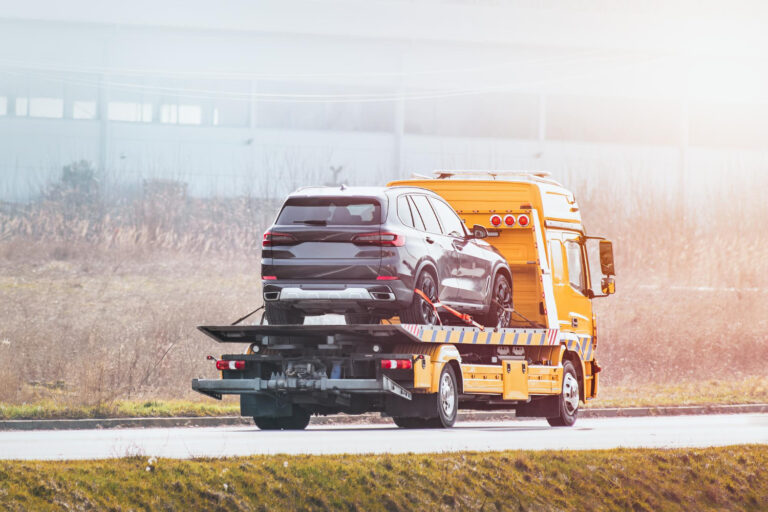Żółta ciężarówka holownicza przewozi czarnego SUV-a na platformie wzdłuż drogi, z rozmytą trawą i budynkami w tle w jasny, mglisty dzień.