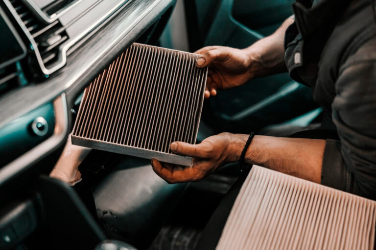 Osoba z lekko zabrudzonymi rękami trzyma prostokątny kabinowy filtr powietrza wewnątrz samochodu, przygotowując się do jego montażu lub demontażu z obszaru schowka na rękawiczki.