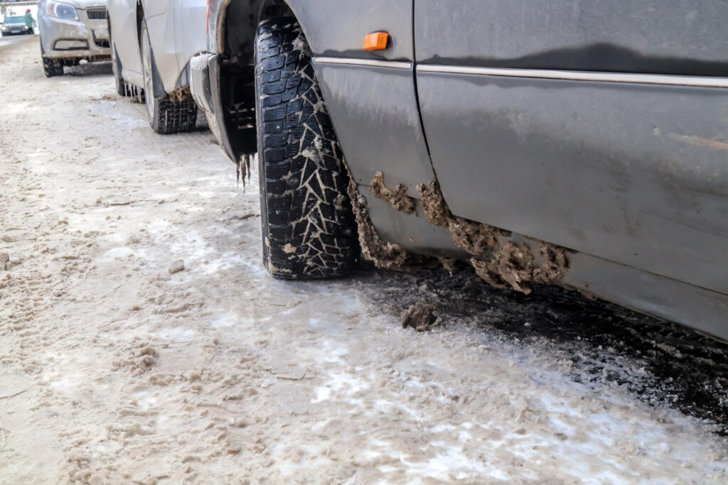 Zbliżenie samochodu zaparkowanego na zaśnieżonej, oblodzonej ulicy pokazuje błoto i lód gromadzące się na kole i dolnej części, z innymi samochodami zaparkowanymi w tle.