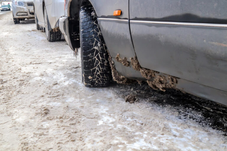 Zbliżenie samochodu zaparkowanego na zaśnieżonej, oblodzonej ulicy pokazuje błoto i lód gromadzące się na kole i dolnej części, z innymi samochodami zaparkowanymi w tle.