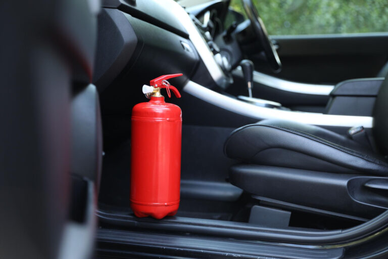 Czerwona gaśnica umieszczona na podłodze przed siedzeniem pasażera w samochodzie z czarnym wnętrzem.