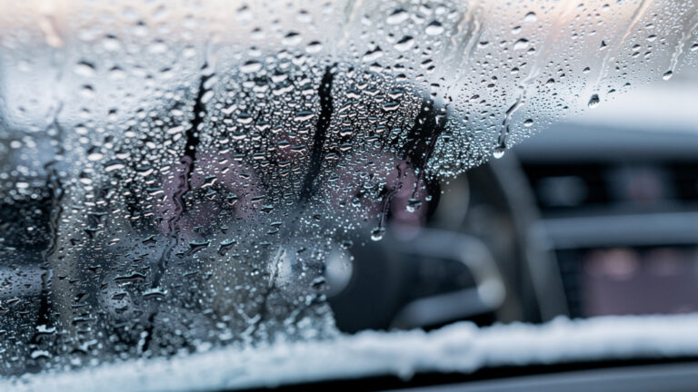 Zbliżenie przedniej szyby samochodu częściowo oczyszczonej z kondensatu i kropelek wody, odsłaniającej rozmazany widok deski rozdzielczej i kierownicy wewnątrz samochodu.